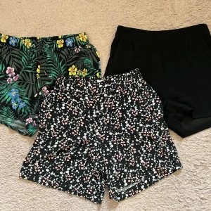 Svarta shorts med blommigt mönster - Säljer tre par shorts, ett med blommigt mönster i vitt och rosa, en helsvart och en i grönt mönster. Två av shortsen har elastisk midja och avslappnad passform, perfekta för varma dagar. Tredje shortsen har ett blixtlås på baksidan. Materialet känns mjukt och luftigt, troligen bomull eller polyester. Supersnygga till sommaren!