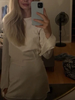 Vit kortklänning med öppen rygg - Säljer en stilren vit kortklänning med långa ärmar och en snygg öppen rygg. Klänningen  är ifrån märket Meshki, och har en enkel, figurnära passform och är tillverkad i ett mjukt, glansigt material som ger en lyxig känsla. Perfekt för dig som vill sticka ut med en elegant look. 