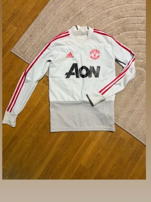 Manchester United träningströja Adidas - Ljusgrå långärmad träningströja från Adidas med Manchester United-logga och AON-tryck på bröstet. Röda Adidas-ränder längs ärmarna och hög krage. Perfekt för fotbollsträning eller som sportig streetwear.