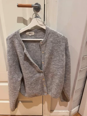 Ljusgrå kofta i ullblandning - Mysig ljusgrå kofta med rund hals och knappar framtill. Tillverkad i en mjuk ullblandning som ger en fluffig känsla. Perfekt att slänga över en t-shirt för en avslappnad look. Klassisk och enkel design som passar till det mesta.