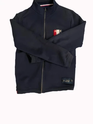 Mörkblå ziptröja från Who’s Ace - Mörkblå ziptröja från Who’s Ace med hög krage och hel dragkedja framtill. Tröjan har ribbade muddar vid ärmslut och nederkant. Perfekt för en avslappnad och sportig stil.