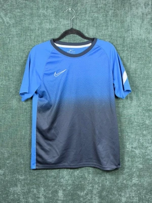Blå Nike Dri-Fit sporttröja - Snygg blå sporttröja från Nike med Dri-Fit material som andas. Tröjan har korta ärmar, rund halsringning och en vit Nike-logga på bröstet. Färgen går från blå upptill till mörkgrå nedtill med ett diskret rutmönster. Perfekt för träning eller match.