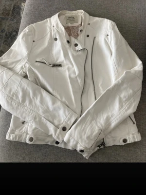 Vit skinnjacka  - Snygg vit skinnjacka från Jofama med coola detaljer som snedställd dragkedja, dekorativa knappar och flera fickor med dragkedja. Jackan har en modern bikerstil och står ut med sin ljusa färg och stilrena design.