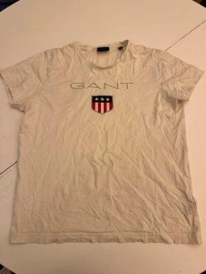 Vit GANT t-shirt med tryck - Vit t-shirt från GANT med klassiskt tryck framtill och ett sköldmärke med stjärnor och ränder. T-shirten har rund halsringning och korta ärmar. Tillverkad i mjuk bomull för en bekväm känsla.
