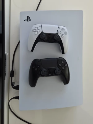 PlayStation 5 med två DualSense-kontroller - PlayStation 5 konsol från Sony i vit färg med två DualSense-kontroller, en vit och en svart. Konsolen har en modern, stilren design och är tillverkad i plast. Perfekt för gaming och upplevelser i nästa generation.