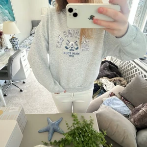 Grå sweatshirt med tryck - Säljer en ljusgrå sweatshirt med blått tryck framtill där det står 'Portland Bay Main Tour 1991' och två korsade åror. Tröjan har rund hals, ribbade muddar och avslappnad passform. Perfekt för en chill och sportig look. Passar S!