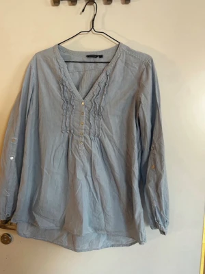 Blå v-ringad blus med volang - Ljusblå blus från H&M med v-ringning, dekorativa volanger och små knappar framtill. Tillverkad i ett tunt, mjukt bomullstyg. Långa ärmar med knappdetaljer som kan vikas upp. Perfekt för en avslappnad men ändå stilsäker look. Rök och djurfritt hem. Pruta gärna !🌸