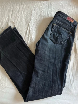 Mörkblå Adriano Goldschmied Jeans - Snygga mörkblå lågmidjade jeans från AG, Adriano Goldschmied med klassisk femficksmodell och kontrastsömmar. Jeansen har raka ben och en ikonisk broderad logga på bakfickan. Tillverkade i slitstark denim med rå kant nedtill för en trendig look.
