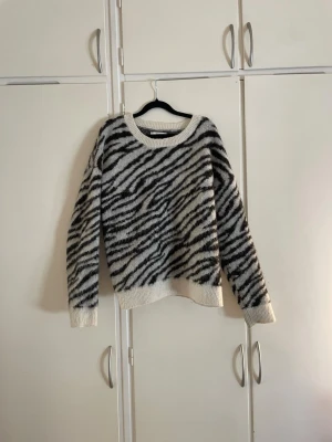 Zebramönstrad stickad tröja - Snygg stickad tröja från Donna i storlek M med zebramönster i svart och vitt. Tröjan har rund halsringning, långa ärmar och ribbstickade muddar vid ärmslut och nederkant. Perfekt för dig som gillar djurmönstrat och vill ha en mysig look. 