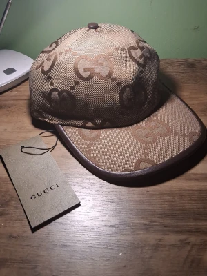 Beige keps med GG-mönster från Gucci - Säljer en beige keps från Gucci med det klassiska GG-monogrammet i brunt över hela kepsen. Kepsen har böjd skärm med bruna kanter och är tillverkad i ett slitstarkt canvasmaterial. Perfekt för dig som vill ha en ikonisk accessoar med lyxig känsla.