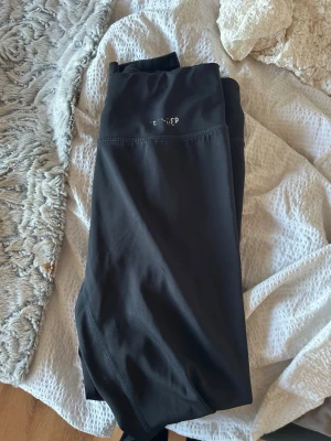 Svarta leggings från STRONGER - Säljer ett par svarta leggings från STRONGER med hög midja och stretchigt material. Perfekta för träning eller chill, med diskret logga framtill. Snygg passform och bekväma sömmar som ger en clean look.
