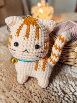 Virkad sur katt nyckelring med bjällra - Supersöt handvirkad nyckelring i form av en beige katt med orange ränder Katten har en liten gul bjällra runt halsen. Dockan virkade med omsorg och noggrann het. Nu är den redo att få en ny ägare.                          Storlek (ungefärlig)                    Huvudomkrets : 17.5 cm                            Höjd : 9 cm 