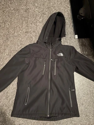 Svart vindjacka från The North Face - Snygg svart vindjacka från The North Face med huva och dragkedja framtill. Jackan har flera praktiska fickor med dragkedja och logga på bröstet och ryggen. Tillverkad i ett slitstarkt, vattenavvisande material som passar perfekt för blåsiga dagar. Jackan är även en gortex jacka så du slipper oroa dig när det regnar. Kontakta mig för fler bilder o tveka inte o höra av dig.
