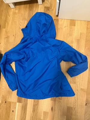 Blå vindjacka med huva från Adidas - Snygg blå vindjacka från Adidas med huva och dragkedja framtill. Jackan har två sidofickor med dragkedja och elastiska muddar vid ärmsluten. Tillverkad i lätt och tåligt syntetmaterial, perfekt för blåsiga dagar. Adidas-logga på ärmen ger en sportig touch.