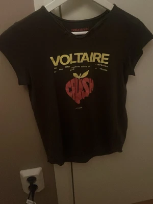 Svart Voltaire t-shirt med tryck - Cool svart t-shirt från Zadig & Voltaire med gul text och ett rött äpple-tryck på framsidan. Baksidan har turnélista med städer i gul text. T-shirten är kortärmad och har en klassisk passform, perfekt för dig som gillar statement-plagg. Den e köpt på nk i stan