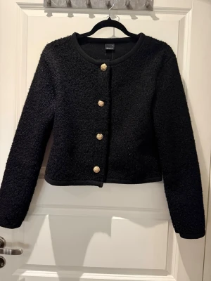 Svart bouclékofta från Gina Tricot - Stilren svart bouclékofta från Gina Tricot med rund halsringning och fyra dekorativa guldfärgade knappar framtill. Kortare modell med lång ärm och mjuk, texturerad yta som ger en trendig look. Koftan är aldrig använd och har prislappen kvar 🌸