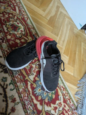 Svarta Nike sneakers med röd detalj - Säljer ett par svarta Nike sneakers med vit sula och röd detalj på hälen. Skorna har mesh-överdel för bra ventilation och klassisk Nike-logga på sidan. Perfekta för dig som vill ha en sportig och stilren look.