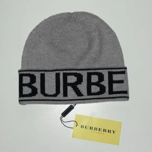 Grå mössa från Burberry - Snygg grå mössa från Burberry med bred uppvikt kant och stor svart logotyptext runt om. Tillverkad i mjukt stickat material som håller dig varm och ger en clean look. Perfekt accessoar för att lyfta din outfit med en exklusiv touch.