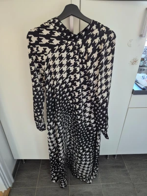 Svartvit långklänning med rosett S - Snygg långklänning i svartvitt rutigt mönster med stor rosett i halsen. Klänningen har långa ärmar och ett klassiskt, trendigt uttryck. Perfekt för dig som vill sticka ut med ett grafiskt mönster och gillar detaljer som knyt i halsen. Helt ny, aldrig använd! Storlek S