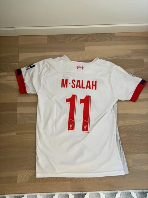 Liverpool bortatröja M. Salah #11 - Säljer en vit Liverpool FC bortatröja från New Balance med röda detaljer och tryck M. Salah samt nummer 11 på ryggen. Tröjan har klubbmärke och sponsorlogga på bröstet samt korta ärmar med röda muddar. Perfekt för dig som älskar fotboll och vill stötta laget.
