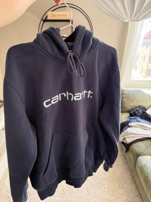 Mörkblå Carhartt hoodie med logga - Snygg mörkblå hoodie från Carhartt med stor vit logga på bröstet. Klassisk känguruficka framtill och justerbar huva med dragsko. Tillverkad i mjukt bomullsmaterial och har ribbade muddar vid ärmar och nederkant. Perfekt för en chill och avslappnad stil.