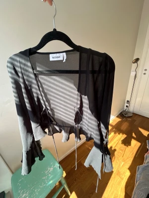 Svart meshkofta från Weekday - Säljer en svart meshkofta från Weekday med volangdetaljer och knytband framtill. Koftan är genomskinlig och har långa ärmar med vågig kant nedtill. Perfekt för lager på lager och en edgy look🪩🥂