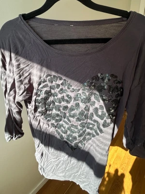 Grå topp med paljett-hjärta - Mjuk grå topp med trekvartsärm och stort hjärta i svarta paljetter på framsidan. Avslappnad passform och rund halsringning. Perfekt för dig som gillar lite extra bling i vardagen🥂💗
