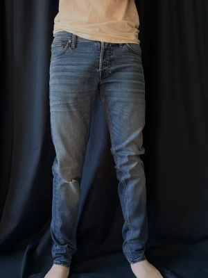 Blå slim fit jeans från Jack & Jones - Snygga blå slim fit jeans från Jack & Jones med klassisk femficksdesign och lätt slitna detaljer på låret. Jeansen har normal midja och raka ben, perfekta till en avslappnad stil. Jeansen är W29 L32