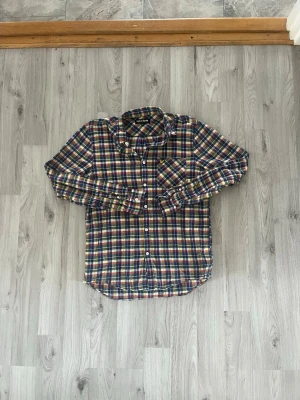 Rutig skjorta i lyle&scott - Klassisk långärmad skjorta från lyle&scottmjuk bomull. Skjortan har ett färgglatt rutmönster i blått, rött, gult och vitt samt knappar framtill och på manschetterna. Perfekt för en avslappnad men ändå stilsäker look.