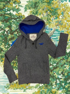  Hollister hoodie stickad - Mysig grå hoodie från Hollister med blått foder i huvan och matchande blå logga på bröstet. Tröjan har känguruficka, dragsko i huvan och en knappdetalj vid halsen. Perfekt för chill dagar och enkel att styla till jeans.