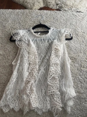 Vit spetsblus med volanger - Säljer en superfin vit ärmlös blus i spets med snygga volangdetaljer framtill och längs axlarna. Blusen har ett vackert mönster och rund halsringning. Storlek S men passar xs också