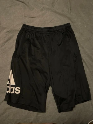 Svarta Adidas shorts - Säljer ett par svarta shorts från Adidas med stor vit logga på höger ben. Shortsen har elastisk midja och sidofickor, tillverkade i ett lätt och snabbtorkande material som passar perfekt till träning eller chill. Klassisk sportig look.