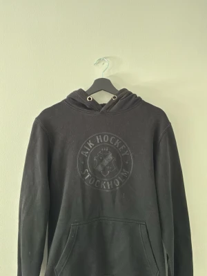 Svart AIK Hockey hoodie - Svart hoodie från AIK Hockey med stor logga på bröstet och känguruficka framtill. Klassisk huva med dragsko och ribbade muddar vid ärmslut och nederkant. Perfekt för dig som vill representera AIK på ett stilrent sätt.