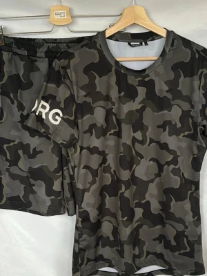 Camouflage t-shirt från Björn Borg - Snygg t-shirt från Björn Borg med grått och svart camouflage-mönster. T-shirten har rund halsringning, korta ärmar och logga på ärmen. Tillverkad i ett lätt och skönt syntetmaterial som passar perfekt för en aktiv livsstil.