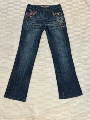 Blå bootcut jeans med brodyr - Snygga blå bootcut jeans med broderade detaljer och nitar på fickorna både fram och bak. Jeansen har en tvättad look och unika mönster. Dom är mid waist. Säljer för att dom är för stora i storleken, det står att dom är i L, men skulle säga M🫶🏼, 