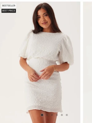 Vit kort klänning med puffärm och knyt - Supersöt vit kort klänning med puffärmar och struktur i tyget. Klänningen har rund halsringning, markerad midja och en snygg öppen rygg med knytband. Perfekt för student 