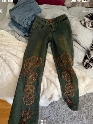 Gröna jeans med broderi från Moschino - Unika gröna jeans från Moschino med färgglada broderier längs båda benen. Jeansen har rak passform, klassisk femficksmodell och detaljer i orange. Materialet är denim i bomull. Perfekta för dig som vill sticka ut med coola detaljer och vintagekänsla.