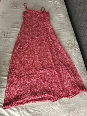 Röd blommig långklänning med smala band - Söt röd långklänning med vita små blommor och smala axelband som knyts. Klänningen har en lätt utställd passform och är perfekt för varma dagar. Materialet känns lätt och luftigt, vilket gör den skön att bära under sommaren. 