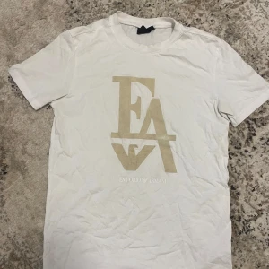 Emporio Armani t-shirt - Säljer en vit t-shirt från Emporio Armani med stort beige EA7-tryck på bröstet. Klassisk rund hals och korta ärmar. Tillverkad i mjuk bomull som känns skön mot huden. Perfekt för dig som gillar stilrena och exklusiva plagg.