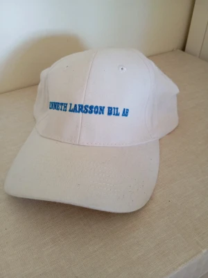 Vit keps med blå text - Säljer en vit keps med böjd skärm och blå broderad text på framsidan där det står 'KENNETH LARSSON BIL AB'. Klassisk modell i bomullstyg med justerbar baksida. Perfekt för dig som gillar enkel och clean stil.