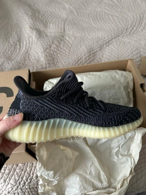 Yeezy Boost 350 V2 svart/gul - Säljer ett par Adidas Yeezy Boost 350 V2 i svart med inslag av grått och en ljusgul, ribbad sula. Ovandelen är i stickad mesh med klassiska Yeezy-detaljer och svarta skosnören. Perfekta för dig som gillar streetwear och vill ha något unikt på fötterna.