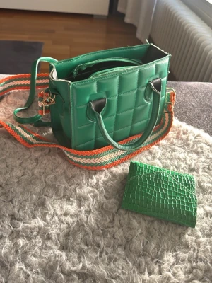 Grön quiltad handväska med axelrem - Snygg grön handväska i quiltad design med två handtag och avtagbar axelrem i grönt, orange och vitt. Väskan är i konstläder och har guldfärgade detaljer. Medföljer matchande grön plånbok i krokopräglat mönster. Perfekt för dig som vill sticka ut. 23x20x8 cm