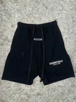 Svarta Essentials Fear of God shorts - Svarta shorts från Essentials Fear of God med vit logga på vänster ben (reflex). De har elastisk midja med snörning och en bakficka. Tillverkade i mjuk bomull, perfekt för chill dagar. Klassisk loose fit och enkel design som funkar till allt.