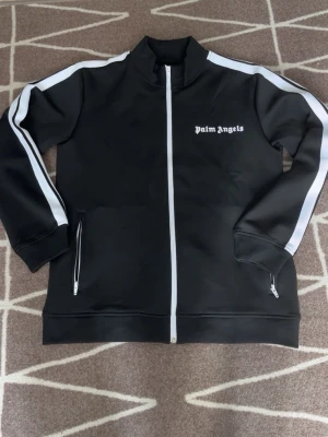 Svart Palm Angels zip hoodie - Snygg svart hoodie från Palm Angels med vit dragkedja och vita ränder längs ärmarna. Märkets logga i vitt på bröstet. Tillverkad i ett glansigt, sportigt material och har två fickor med dragkedja. Perfekt för en streetwear-look. Storlek S men passar till M