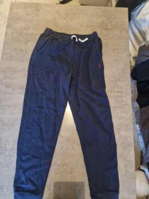 Mörkblå mjukisbyxor Polo Ralph Lauren - Mörkblå mjukisbyxor från Polo Ralph Lauren med dragsko i midjan och fickor på sidorna. Klassisk röd logga broderad på vänster sida. Sköna och avslappnade med resår i bensluten, perfekta för chill dagar. Tillverkade i mjukt material.