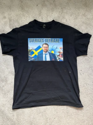 Svart t-shirt med tryck Sveriges Befriare - Svart t-shirt med färgstarkt tryck på bröstet som visar texten 'Sveriges Befriare', en svensk flagga och en blå blomma. T-shirten har rund hals och korta ärmar. Perfekt för dig som vill sticka ut med ett unikt motiv.