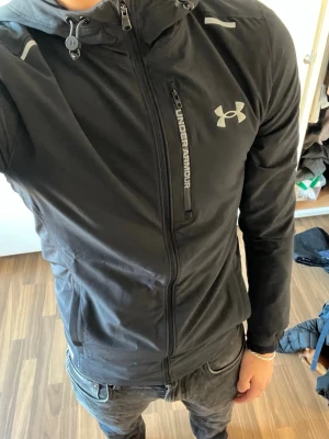 Svart vindjacka från Under Armour - Snygg svart vindjacka från Under Armour med dragkedja framtill, bröstficka med logga och justerbar huva. Jackan har en sportig look med reflekterande detaljer och är tillverkad i ett lätt, vindtätt material som passar perfekt för aktiva dagar.