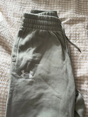 Gröna mjukisbyxor från Under Armour - Snygga gröna mjukisbyxor från Under Armour med resår i midjan och dragsko för perfekt passform. Byxorna har sidofickor och en broderad logga på benet. Tillverkade i mjukt och bekvämt material som passar perfekt för chill eller träning.
