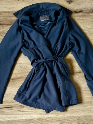 Mörkblå trenchcoat från Vero Moda - Stilren mörkblå trenchcoat från Vero Moda med klassisk krage och knytskärp i midjan. Jackan har långa ärmar och en mjuk, lätt tygkvalitet som ger ett snyggt fall. Perfekt för dig som vill ha en enkel men trendig look. 