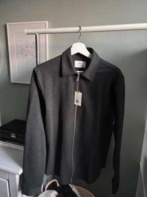 Mörkgrå overshirt från Ciszere - Stilren mörkgrå overshirt från Ciszere med rak passform och klassisk krage. Dragkedja framtill och diskret struktur i tyget. Perfekt lager-på-lager-plagg för en clean och modern look. Materialet känns mjukt och följsamt.